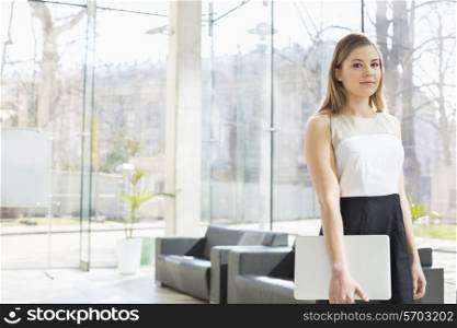 Portrait of confident businesswoman holding laptop while standing at office lobby
