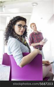 Portrait of confident businesswoman sitting on chair with colleague standing in background at creative office