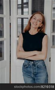 Portrait of confident elegant young woman with her arms crossed. Redhead model in casual outfit is standing and smiling near door. Luxurious apartment interior. Concept of style, fashion and beauty.. Portrait of confident elegant young woman with her arms crossed. Model is standing and smiling.