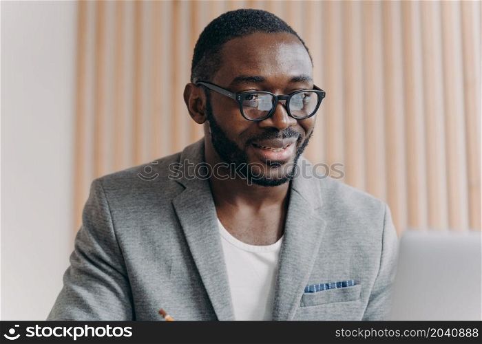 Portrait of confident handsome young african businessman in formal wear sitting at desk in front of laptop in office, looking at computer screen, surfing internet. Diverse business people concept. Handsome young african businessman in formal wear sitting at desk in front of laptop in office,