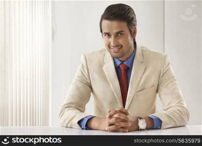 Portrait of confident Indian businessman with hands clasped at office desk