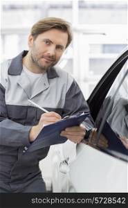 Portrait of confident mechanic holding clipboard while leaning on car&rsquo;s window in workshop