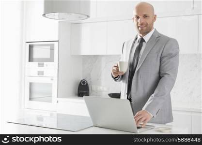 Portrait of confident mid adult businessman having coffee while using laptop in kitchen