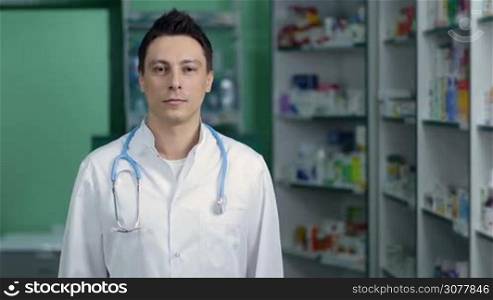 Portrait of confident pharmacist with stethoscope in white lab coat standing with crossed arms in pharmacy and smiling. Medical doctor man with folded crossed hands posing with happy toothy smile on drugstore background. Health care and pharmacology.