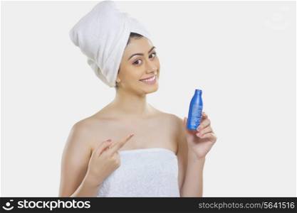 Portrait of confident woman wrapped in towel showing hair oil bottle against white background