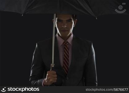 Portrait of confident young businessman holding umbrella against black background