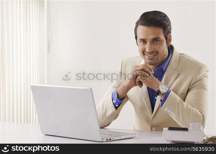 Portrait of confident young businessman with laptop sitting at office desk