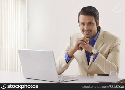 Portrait of confident young businessman with laptop sitting at office desk
