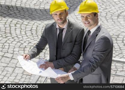Portrait of confident young businessmen in hard hats examining blueprint outdoors