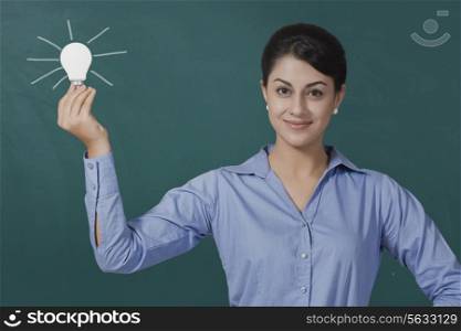 Portrait of confident young businesswoman holding light bulb against green board at office