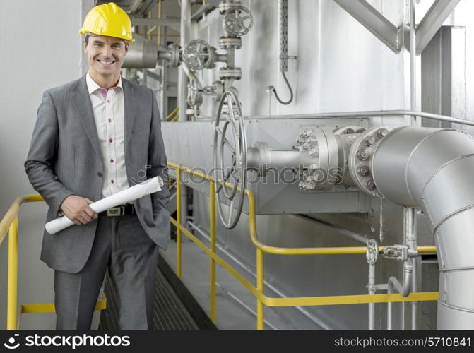 Portrait of confident young male architect holding blueprint by machinery in industry