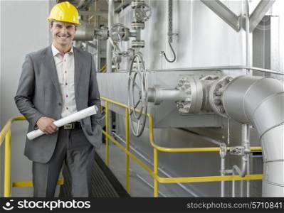 Portrait of confident young male architect holding blueprint by machinery in industry