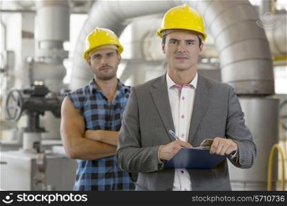 Portrait of confident young manager writing on clipboard with manual worker in background at industry