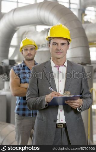 Portrait of confident young manager writing on clipboard with manual worker in background at industry