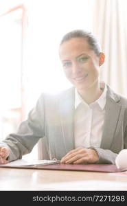 Portrait of confident young saleswoman sitting with document at table against window in apartment