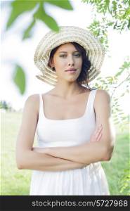 Portrait of confident young woman in sundress and hat standing arms crossed in park