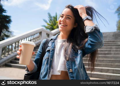 Portrait of confident young woman smiling and holding a cup of coffee while walking outdoors. Urban concept.