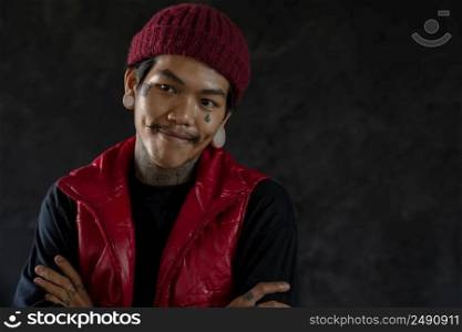 Portrait of cool Asian hip hop young bearded man with tattoo on face, neck and hands standing  smiling and cross his arms with big ear piercing and wear red knit hat and vest. Black background