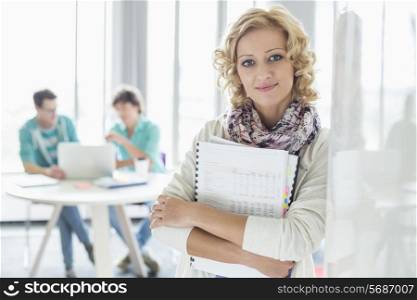 Portrait of creative businesswoman holding files with colleagues working in background at office