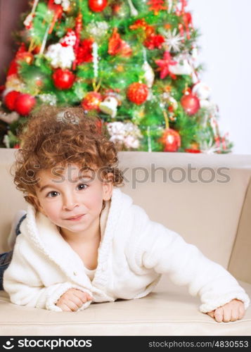 Portrait of cute adorable little boy lying down on the couch near beautiful decorated Christmas tree, spending winter holidays at home