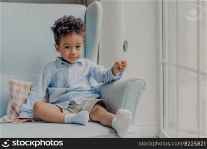 Portrait of cute adorable little mulatto boy with big eyes holding round blue sweet and tasty lollipop and looking at camera, casually dressed sitting alone on comfy armchair in minimalist room. Cute little african american boy with lollipop indoors