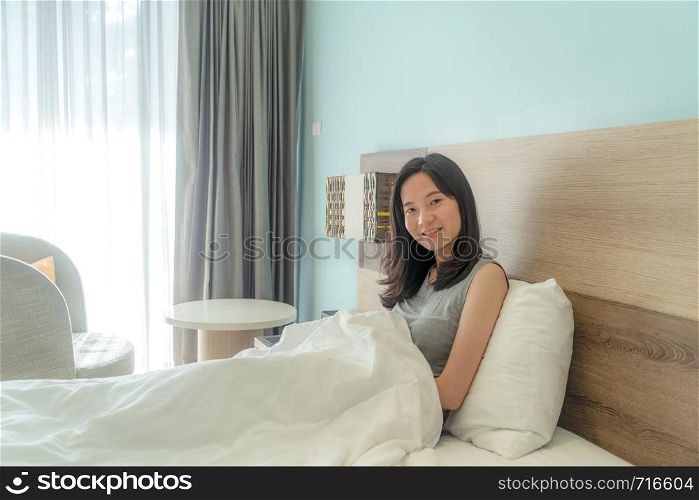 Portrait of cute Asian woman looking at camera on bed in a modern bedroom with white blanket.