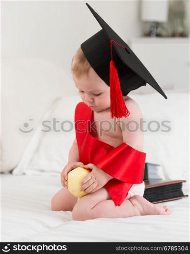 Portrait of cute baby boy in graduation cap holding apple. Concept of baby education