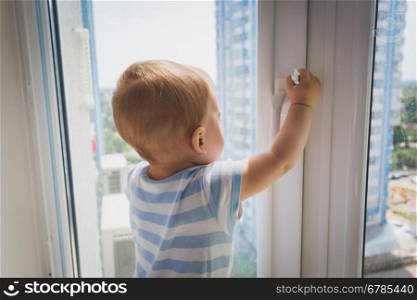 Portrait of cute baby boy pulling by the window handle. Concept of child in danger