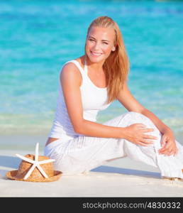 Portrait of cute blond girl sitting on white sandy beach on blue water background, happy summer vacation, travel and tourism concept