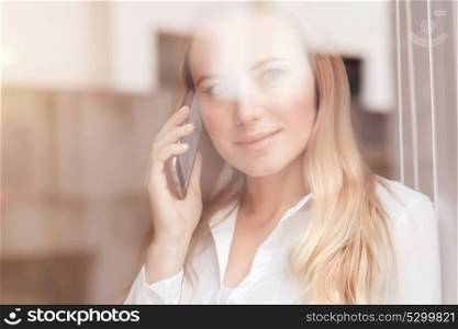 Portrait of cute blond woman standing near window at the office and talking on the phone, mobile connection, lifestyle of modern people