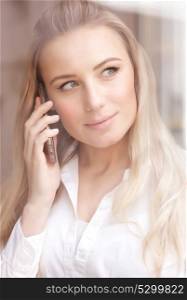 Portrait of cute blond woman standing near window at the office and talking on the phone, mobile connection, lifestyle of modern people