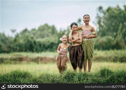 Portrait of cute boy shirtless crossed arms and two lovely girls in Thai traditional dress put beautiful flower on her ear standing in rice field, laughing and look at camera, copy space