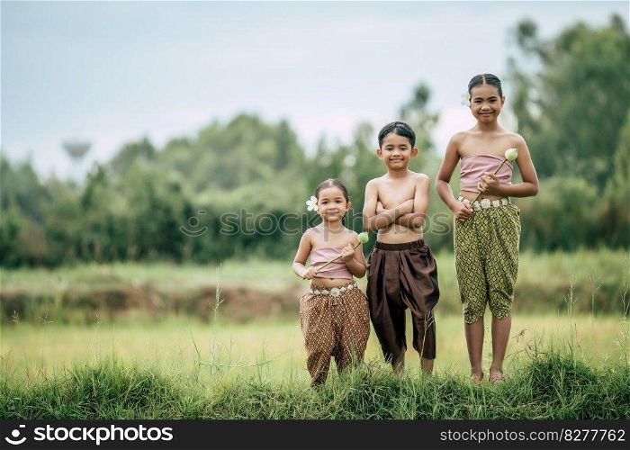 Portrait of cute boy shirtless crossed arms and two lovely girls in Thai traditional dress put beautiful flower on her ear standing in rice field, smile and looking at camera, copy space