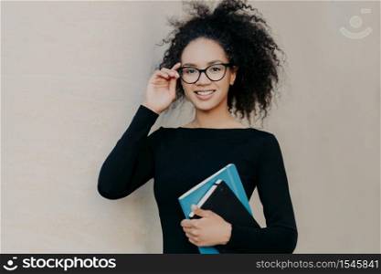 Portrait of cute frizzy young woman with gentle smile, keeps hand on rim of glasses, wears casual black jumper, holds notepad for writing notes, isolated on grey background, studies at college