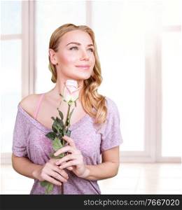 Portrait of cute gentle woman standing near window at home and holding in hands fresh rose, romantic gift on Valentine’s day