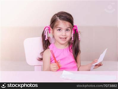 Portrait of Cute girl cuts paper with a red scissors while sitting at pink table. Selective focus and small depth of field.. Portrait of Cute girl cuts paper with a red scissors