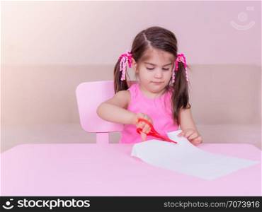 Portrait of Cute girl cuts paper with a red scissors while sitting at pink table. Selective focus and small depth of field.. Portrait of Cute girl cuts paper with a red scissors