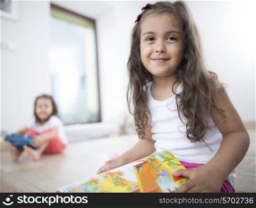 Portrait of cute girl holding book with sister in background at home