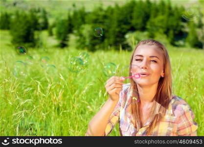Portrait of cute happy woman having fun outdoors, sitting on fresh green grass field and with pleasure blowing soap bubbles, enjoying life