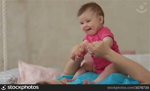 Portrait of cute infant girl laughing while sitting on her mother&acute;s chest in modern bedroom. Happy young mother playing with her cute little daughter in bed while relaxing at home. Family enjoying time together in domestic interior.