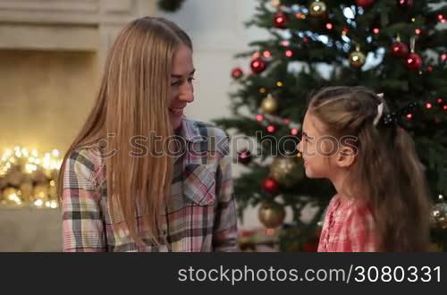 Portrait of cute little girl kissing her beautiful mother at Christmas. Loving daughter giving a kiss to her smiling mom during family xmas eve celebration together in decorated living room. Dolly shot.