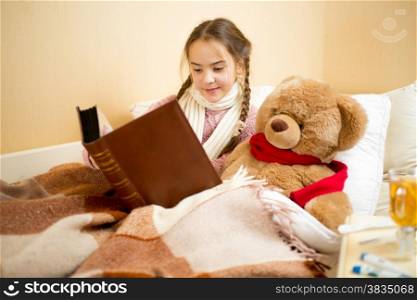 Portrait of cute little girl lying in bed and reading book to teddy bear