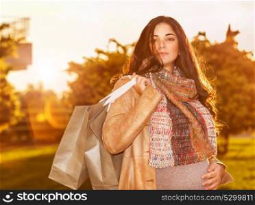 Portrait of cute pregnant woman with shopping bags standing on backyard in warm autumn sunny day, preparing to child birth