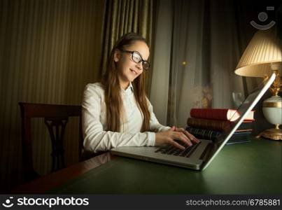 Portrait of cute schoolgirl in white shirt doing homework at laptop in dark room
