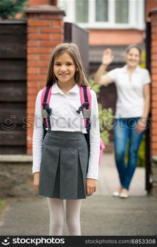 Portrait of cute schoolgirl standing in front of house. Mother waving standing in doorway