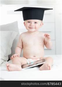 Portrait of cute smiling baby boy in graduation cap sitting on sofa with tablet pc