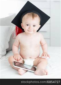 Portrait of cute smiling baby in graduation cap sitting on sofa and holding digital tablet. Concept of smart baby.
