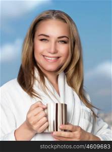 Portrait of cute smiling girl drinking tea on the balcony over blue sky background, wearing white bathrobe and having breakfast, with pleasure relaxing at home