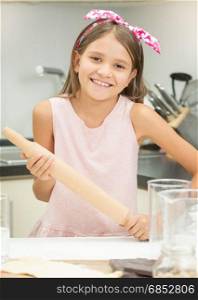 Portrait of cute smiling girl holding wooden rolling pin while making dough