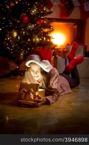 Portrait of cute smiling girl lying on floor at fireplace and looking inside of glowing gift box
